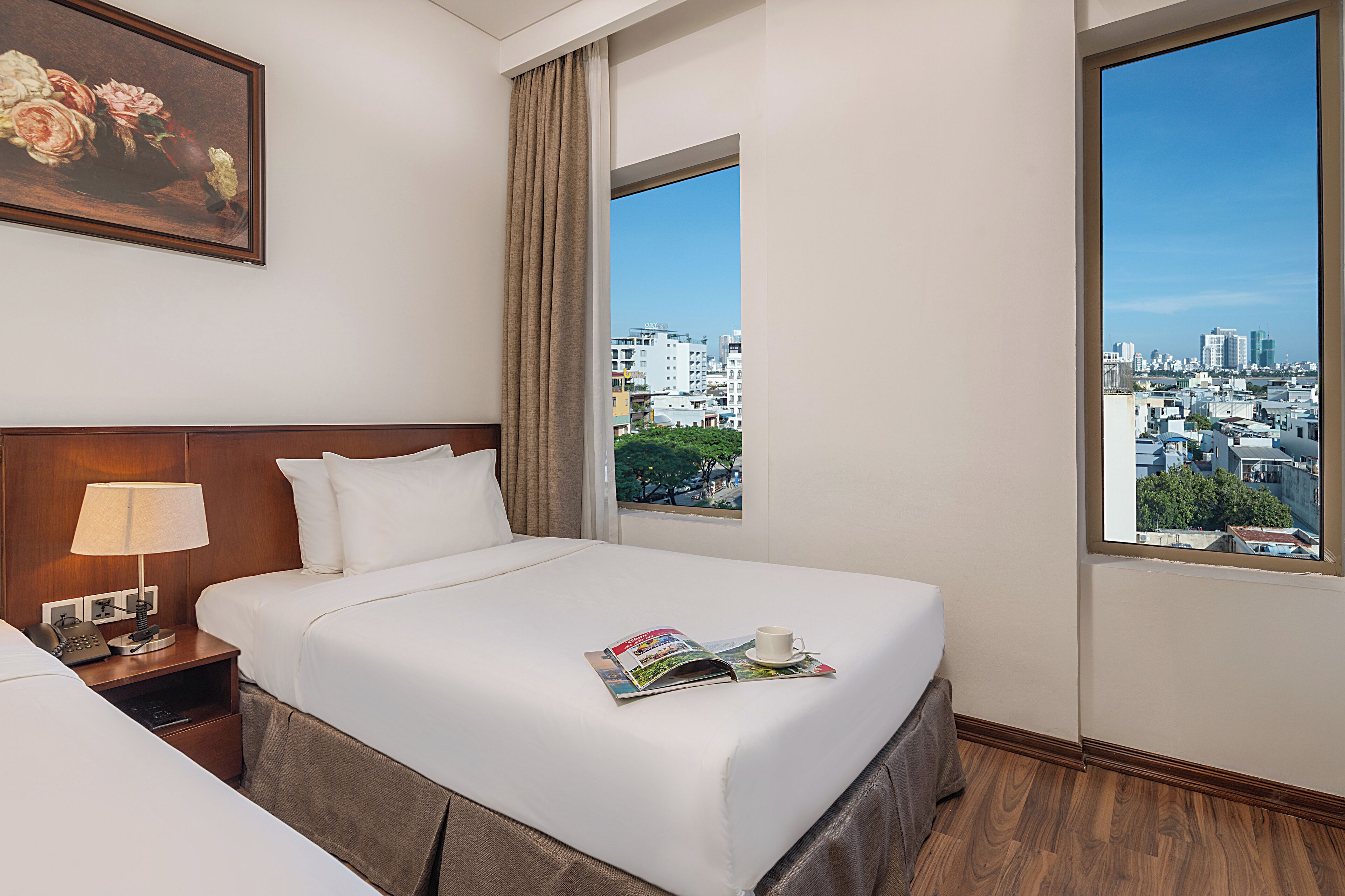 Habitación de hotel con dos camas, una mesita de noche con lámpara, una revista sobre la cama y vista a la ciudad por la ventana.