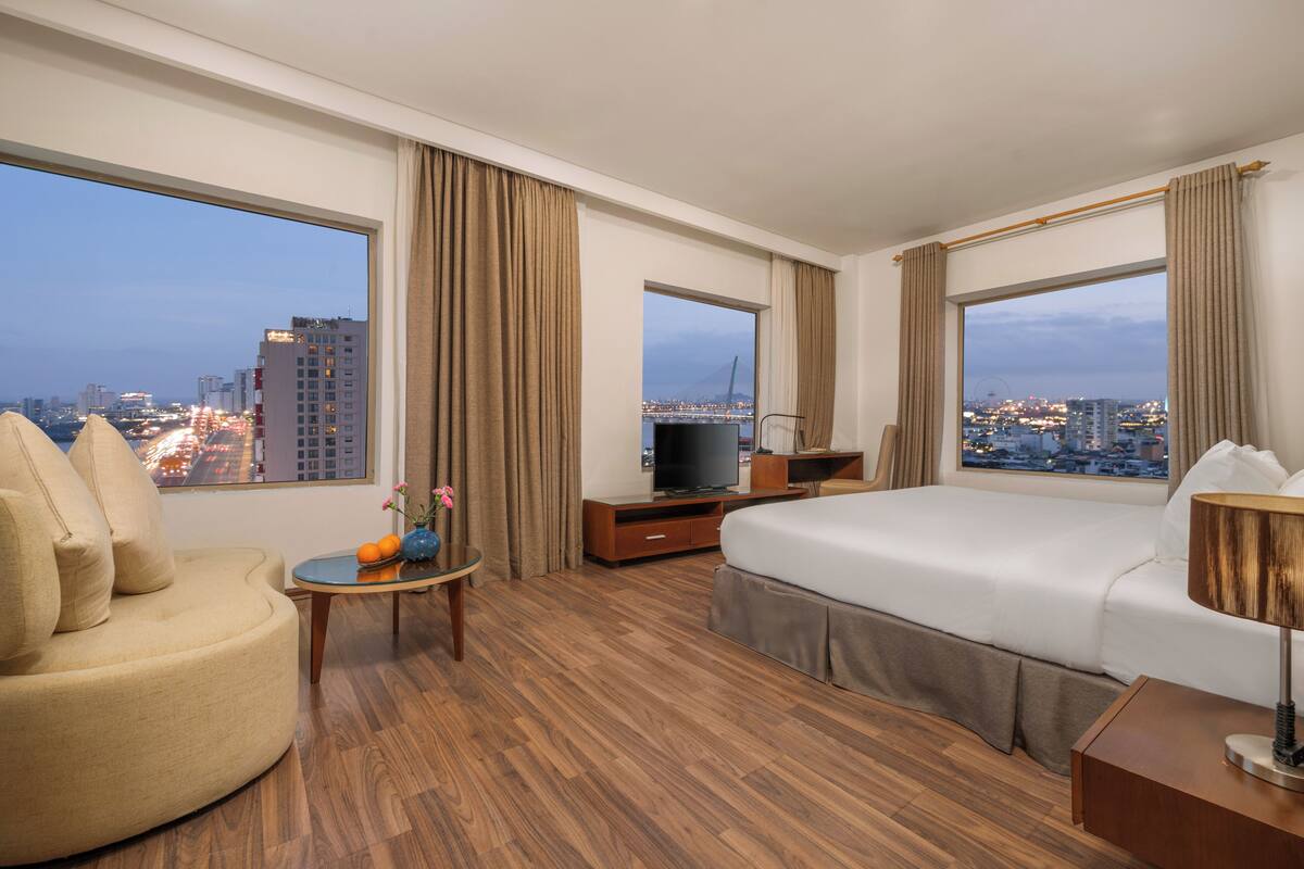 A hotel room with a large bed, a sofa, a TV, and a view of the cityscape.
