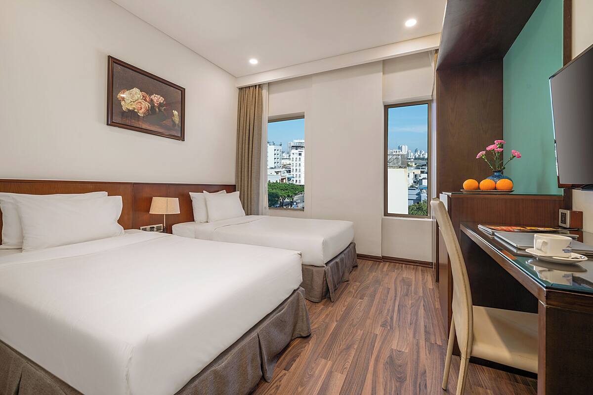 A hotel room with two beds, a desk, a chair, a TV, a window with a city view, and a framed picture on the wall.
