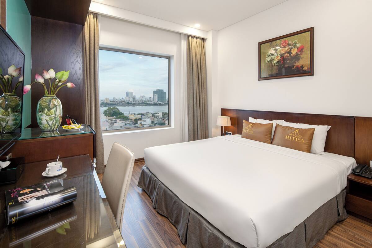 A hotel room with a large bed, a desk with a cup and saucer, a vase with flowers, and a painting on the wall.