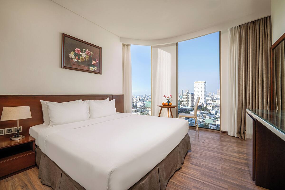 A hotel room with a large bed, a nightstand with a lamp, a small table with a vase of flowers, and a view of the city through large windows.