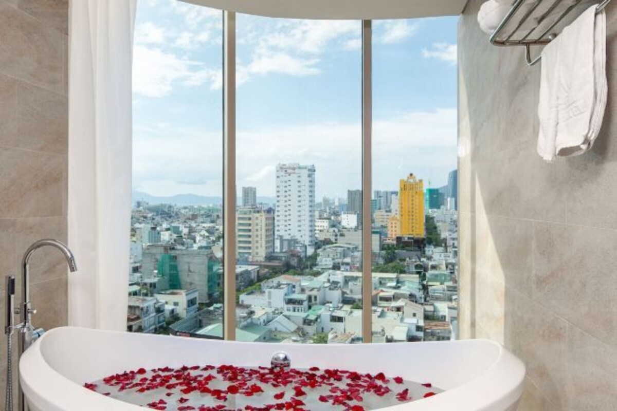 A modern bathroom with a bathtub filled with rose petals, a large window offering a cityscape view, and a towel rack on the wall.
