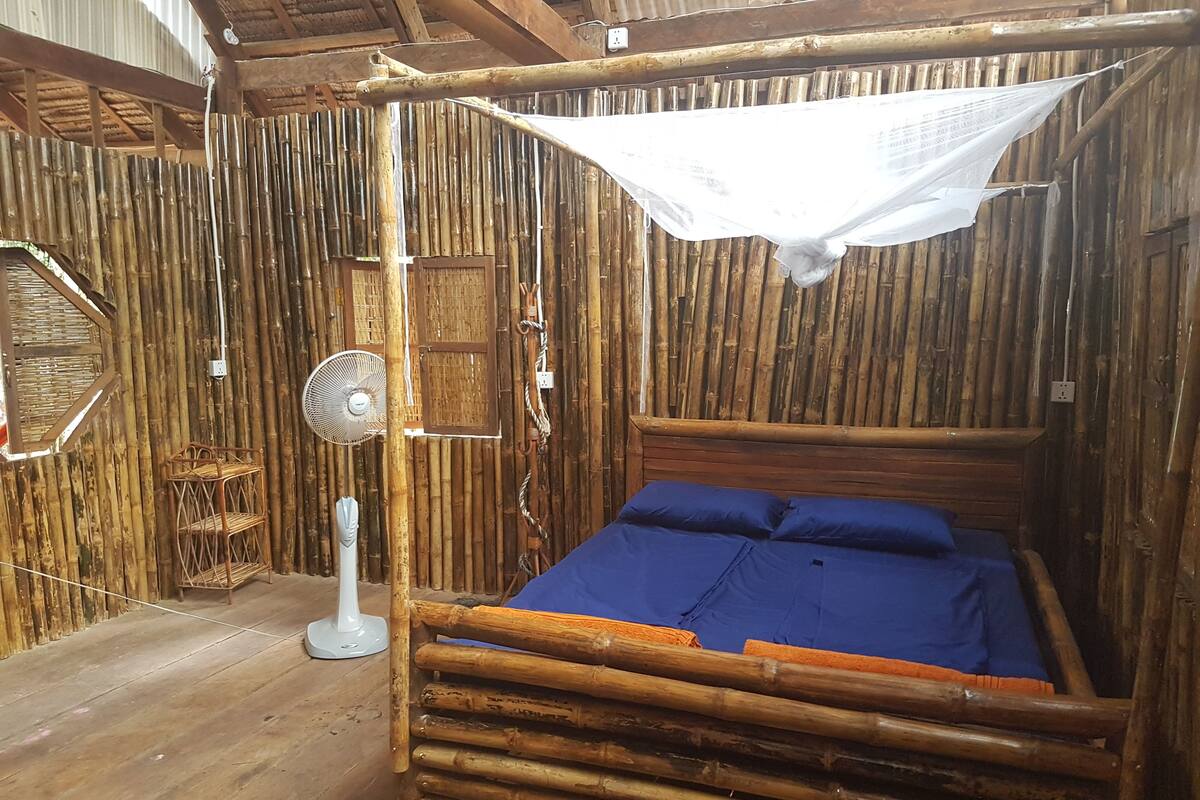 Una habitación con paredes de bambú, una cama, un ventilador y una mosquitera.