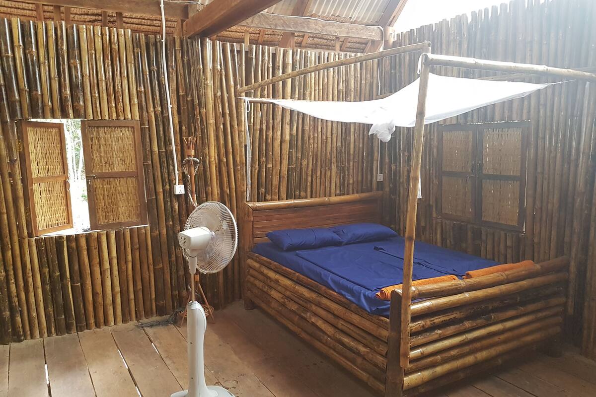 Una habitación con pared de bambú, una cama con dosel y un ventilador de pie.