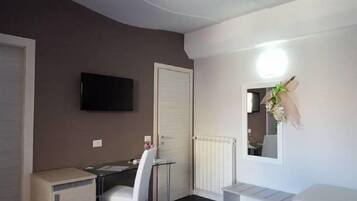 A room with a flat-screen TV mounted on the wall, a white radiator, a small table with a chair, and a white door.