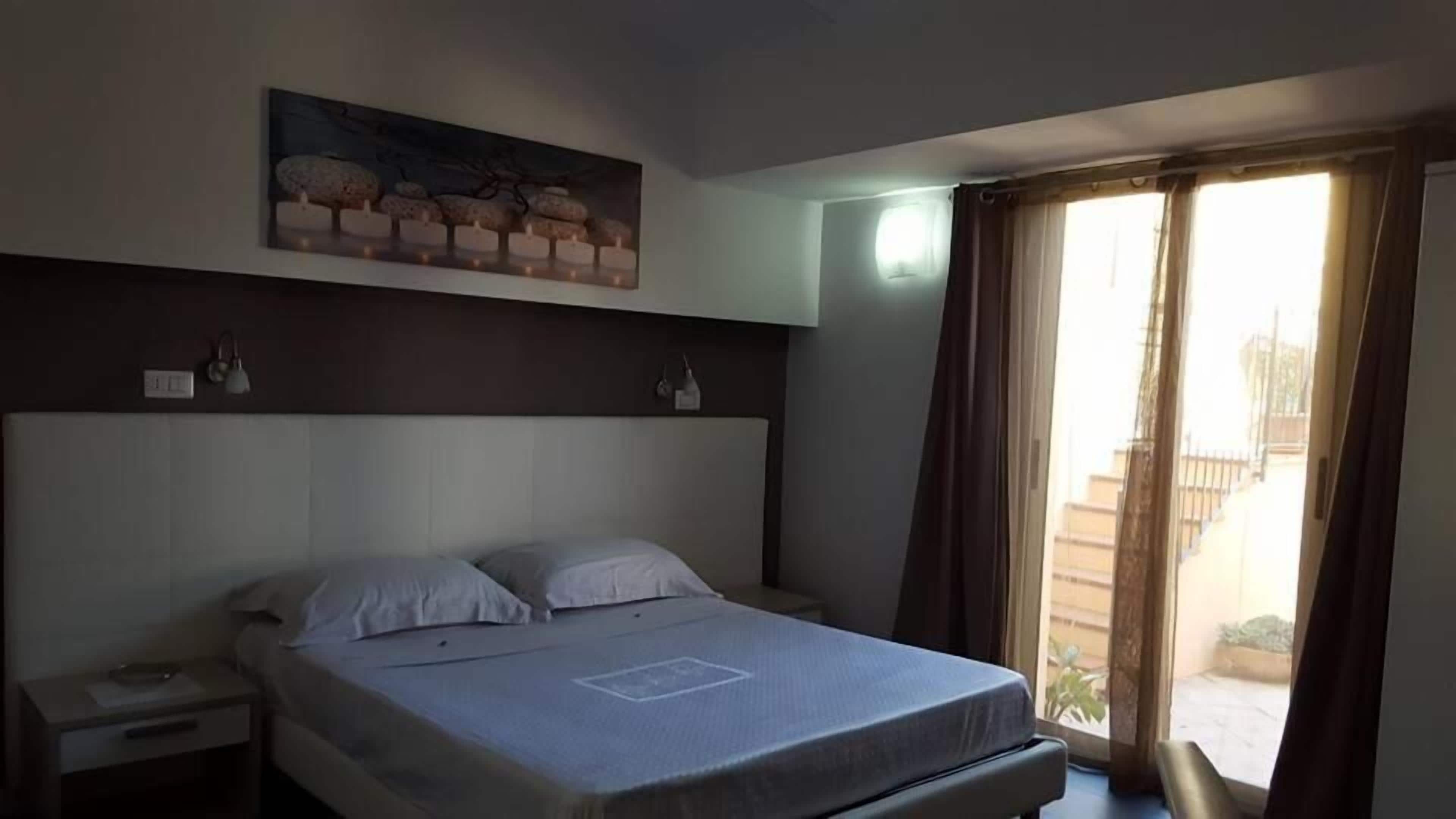 Habitación de hotel con una cama grande, una mesita de noche, un cuadro sobre la cama y una ventana con cortinas.