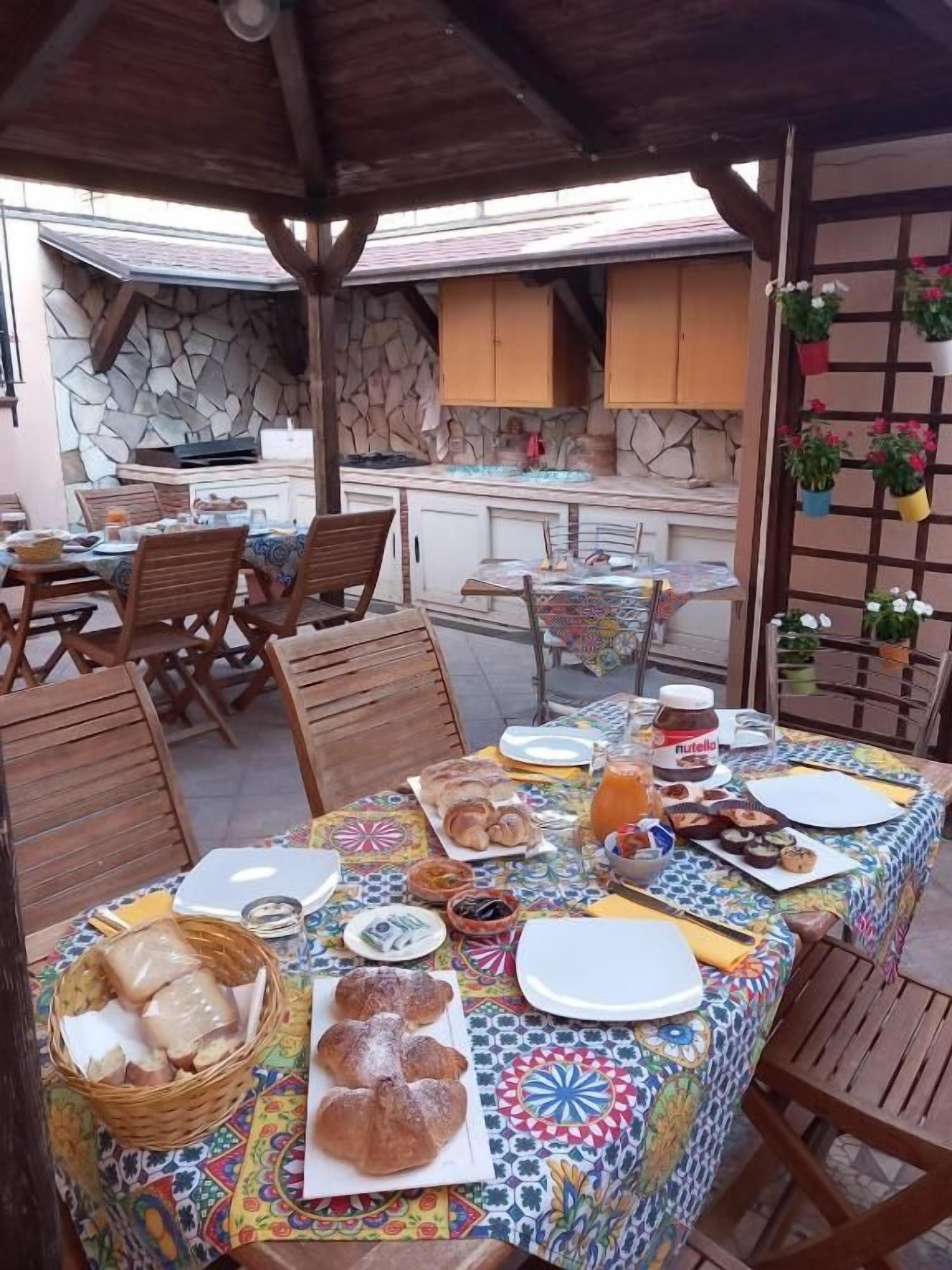 Zona pranzo all'aperto con patio coperto, parete in pietra, sedie di legno, tavolo con tovaglia colorata e angolo cottura sullo sfondo.