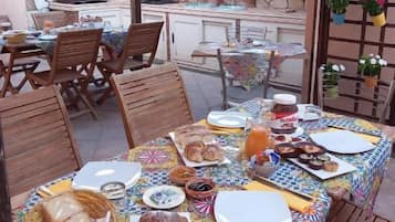 Zona pranzo all'aperto con patio coperto, parete in pietra, sedie di legno, tavolo con tovaglia colorata e angolo cottura sullo sfondo.