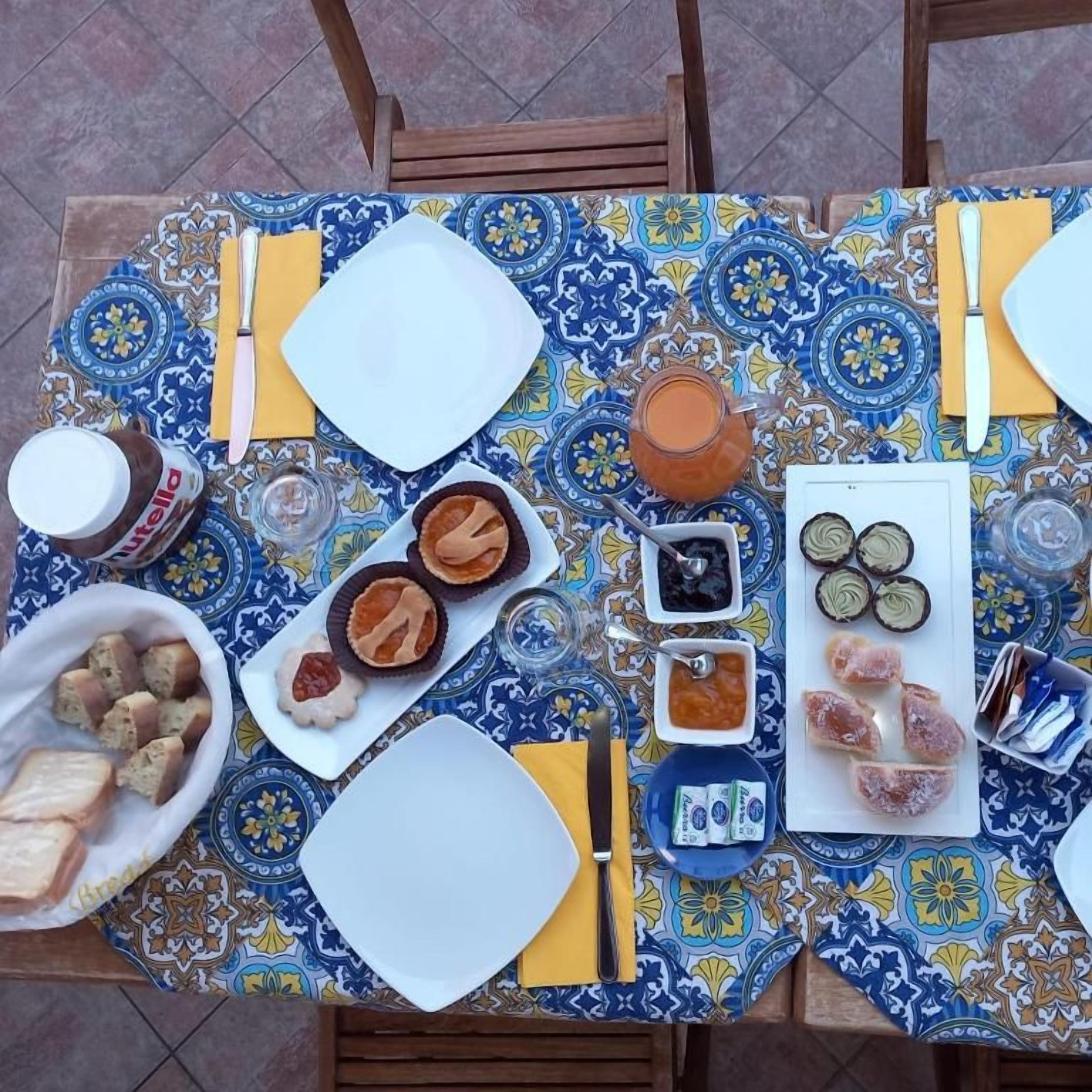 A table set with various food items and condiments, including bread, pastries, and a jug of juice, on a patterned tablecloth.