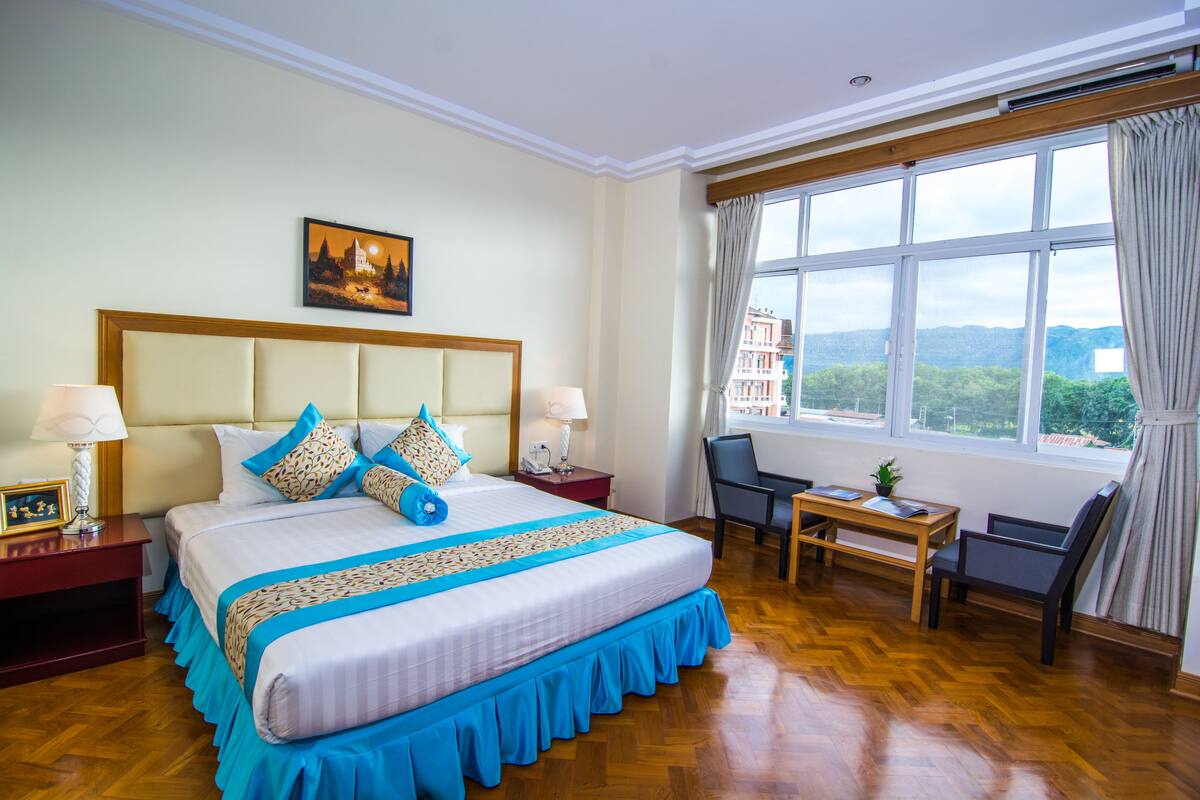 A hotel room with a large bed, a desk, and a chair. There is a window with a view of buildings and trees.