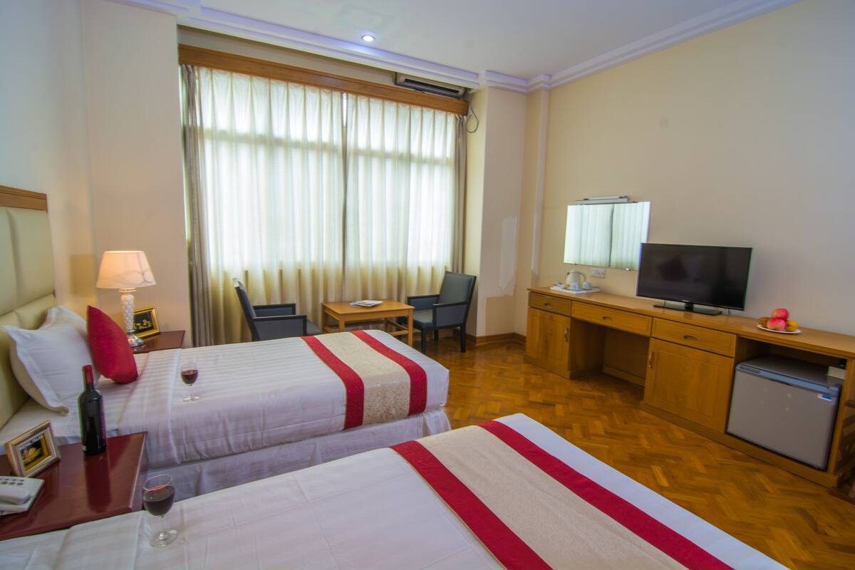 A hotel room with two beds, a desk with a television, a small kitchenette, and a window with curtains.