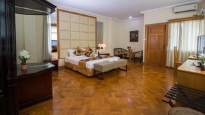 A hotel room with a wooden floor, a bed with a beige headboard, a desk with a computer, a chair, and a small table with a lamp. - Royal Inlay Hotel (Nyaungshwe)