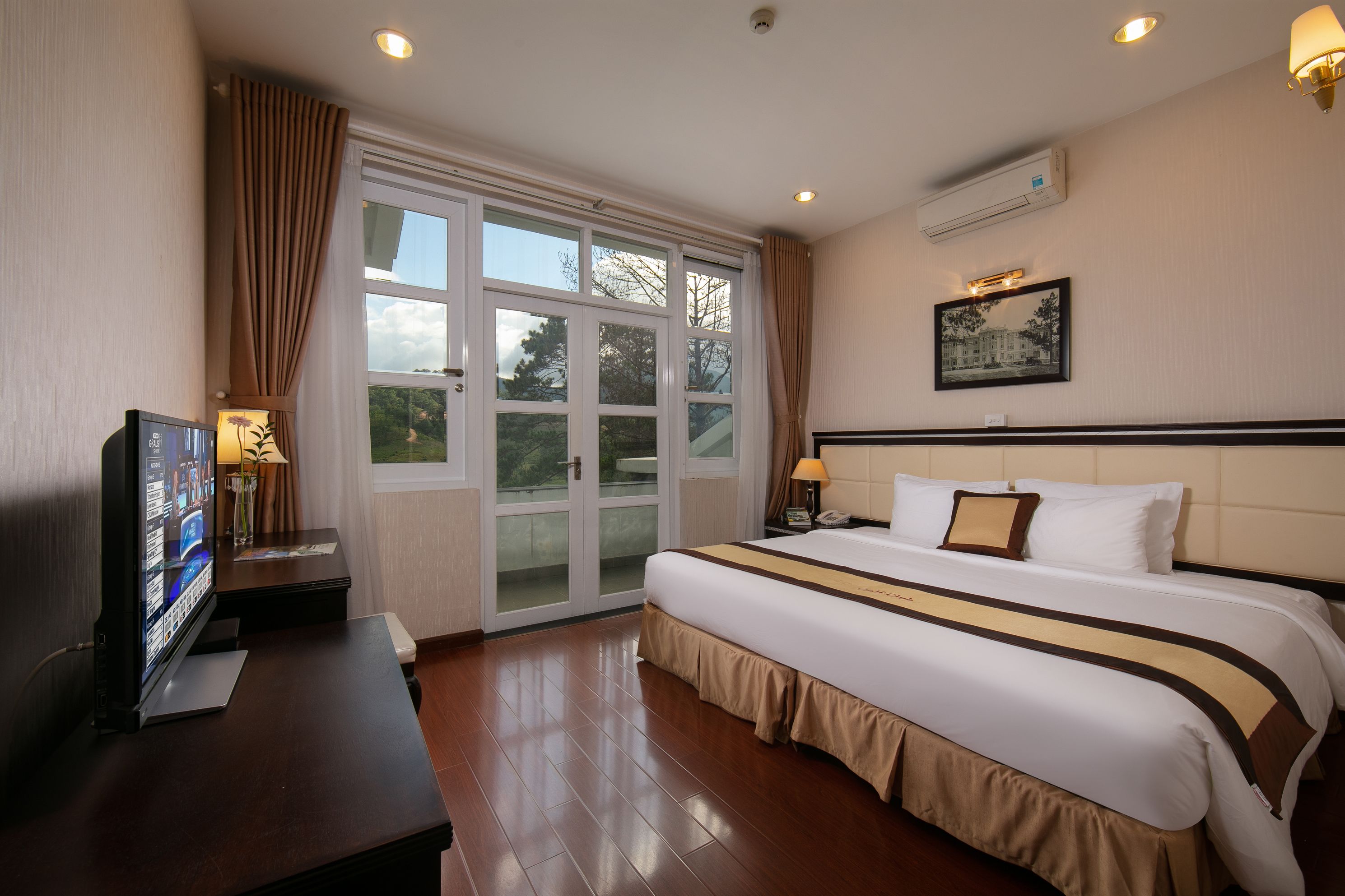 A hotel room with a large bed, a desk with a television, a window with curtains, and an air conditioning unit on the wall.