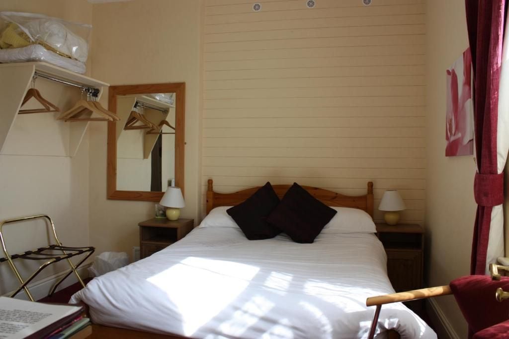 Une chambre d’hôtel comprenant un lit, une tête de lit en bois, une table de chevet avec une lampe, un miroir, une armoire avec des cintres et une fenêtre avec des rideaux.