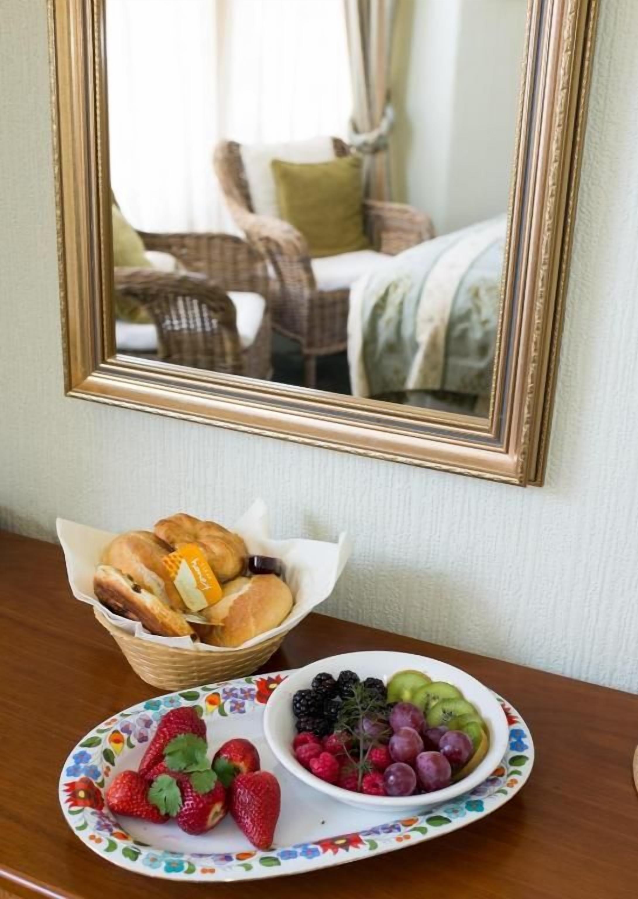 Una colazione con una sedia in vimini, un letto con un cuscino verde, un cestino di panini e un piatto con fragole e frutti di bosco.