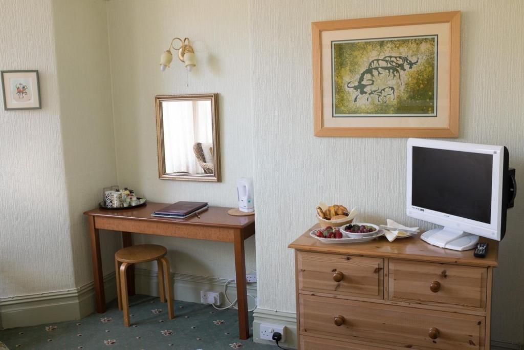 A room with a desk, a chair, a dresser with drawers, a television, a framed picture on the wall, and a small table with a basket and a plate of fruit.