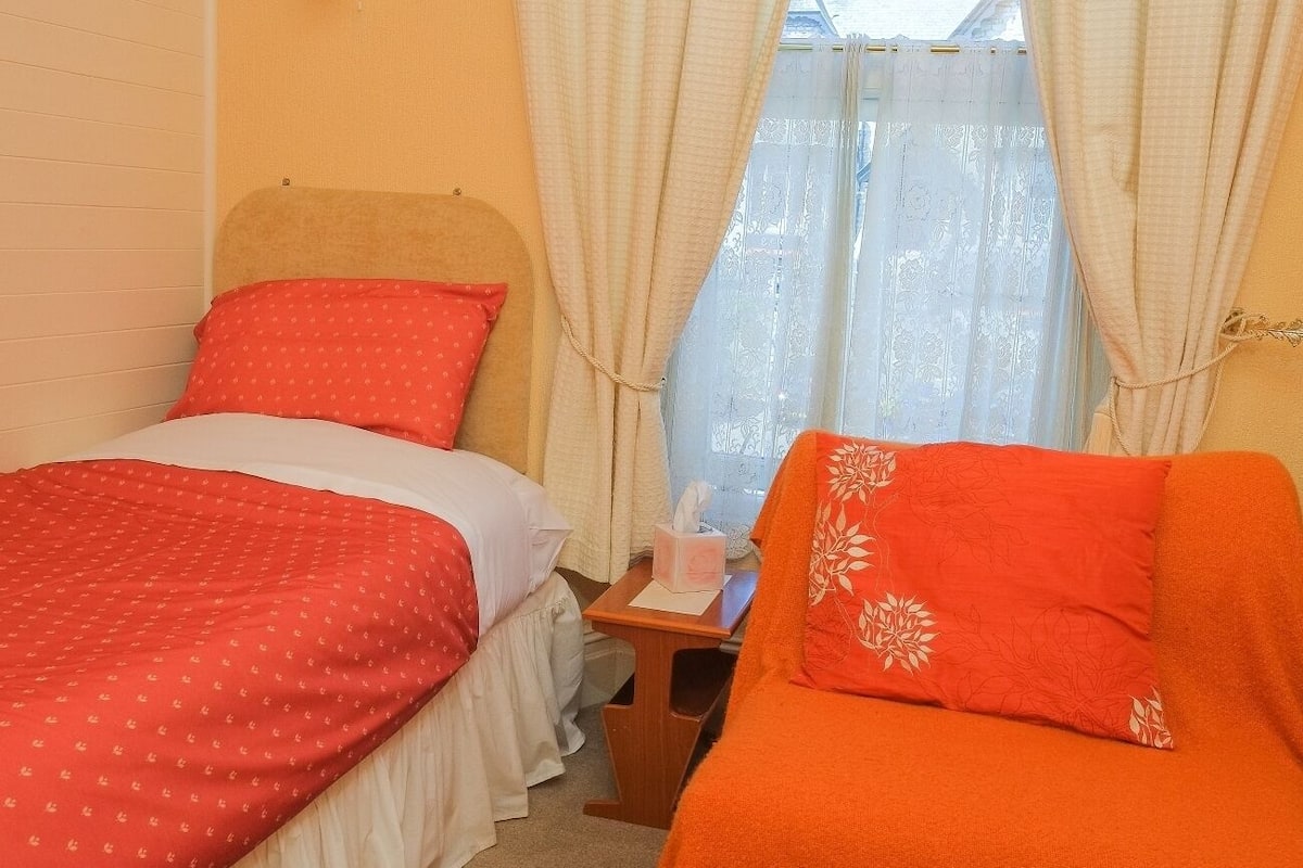 A room with two beds, one with red bedding and the other with white bedding. There is a small wooden table with a tissue box and a bottle. The room has orange walls and a window with sheer curtains.