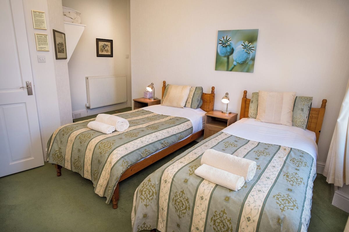 A room with two beds, each with a wooden headboard, a radiator, and a framed picture on the wall.