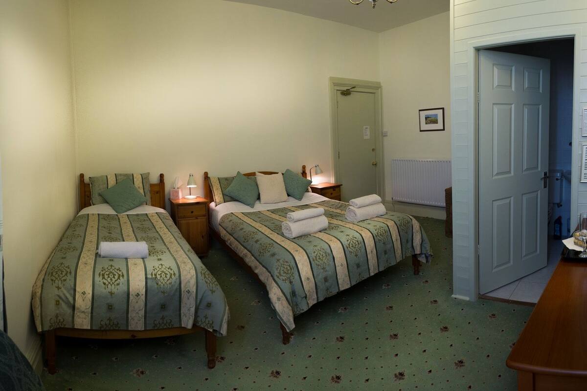 A bedroom with two single beds, each with a bedside table and lamp, a radiator, and a door leading to another room.