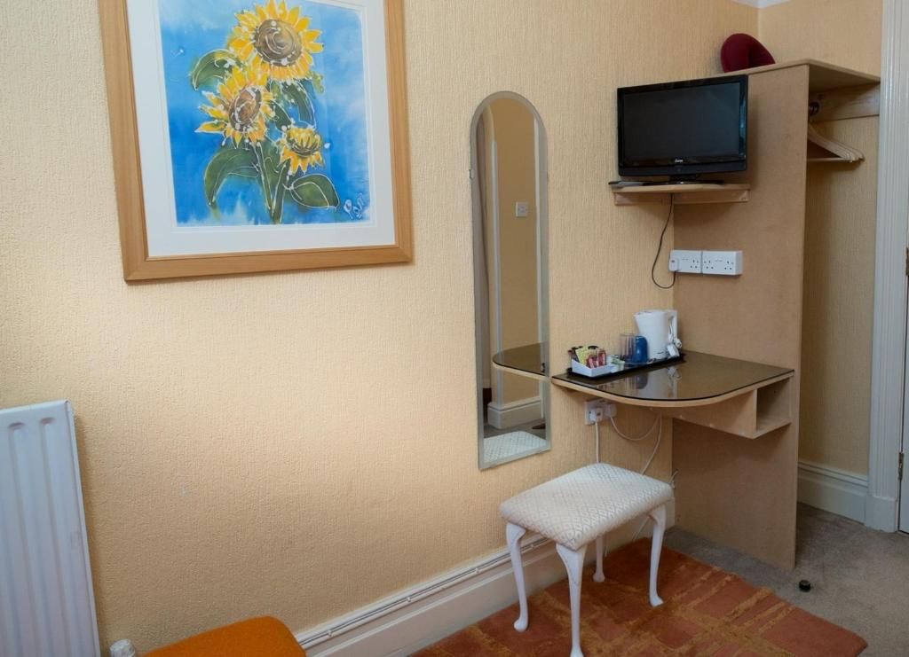 A room with a framed painting of sunflowers, a mirror, a television on a shelf, a small white bench, and a radiator.