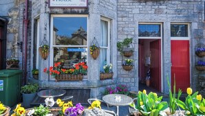 A stone building with a sign reading "Somerset House" and a "NO VACANCIES" sign. There are flower baskets on the windowsill and a small outdoor seating area with tables and chairs. - Somerset House (Grange-over-Sands)