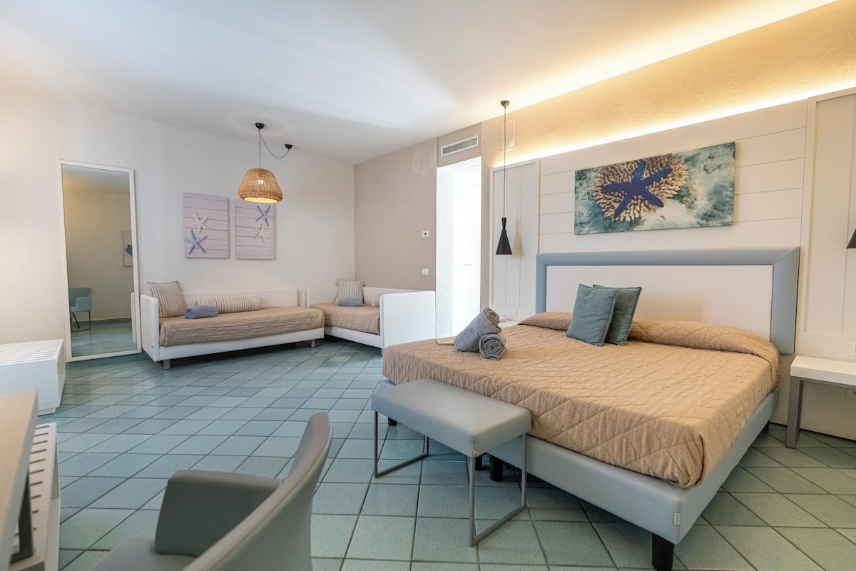 A modern bedroom with a large bed, a smaller sofa, a bench, and a piano.