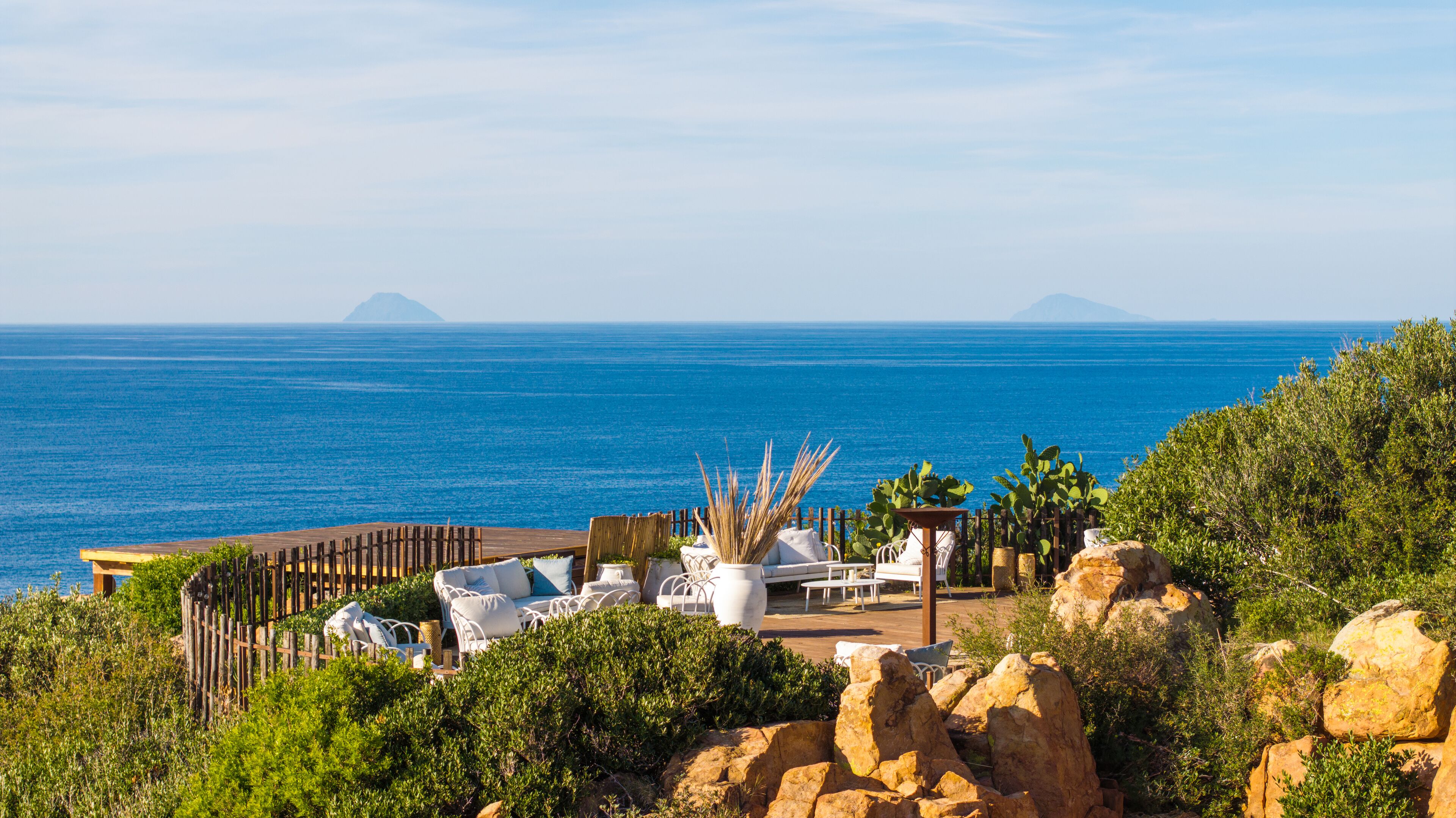 A coastal outdoor seating area with white wicker furniture, a wooden deck, and a view of the sea and distant mountains.