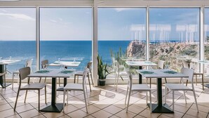 Un restaurante con amplios ventanales que ofrecen vista al océano y a una costa rocosa. Las mesas están dispuestas con sillas blancas y manteles individuales verdes.