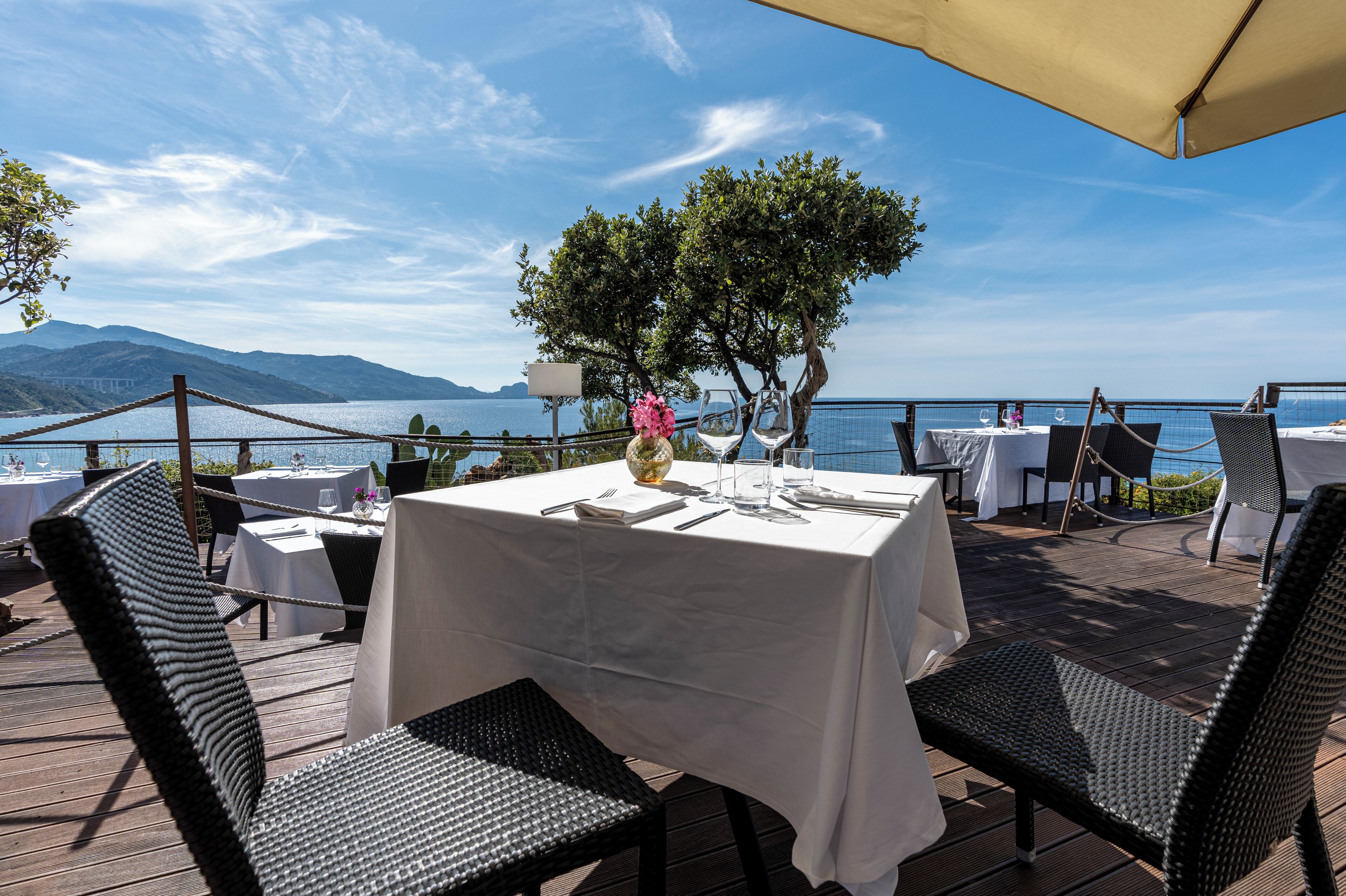 Un espace de restauration en plein air avec des tables pour deux, offrant une vue sur un plan d’eau et des montagnes.