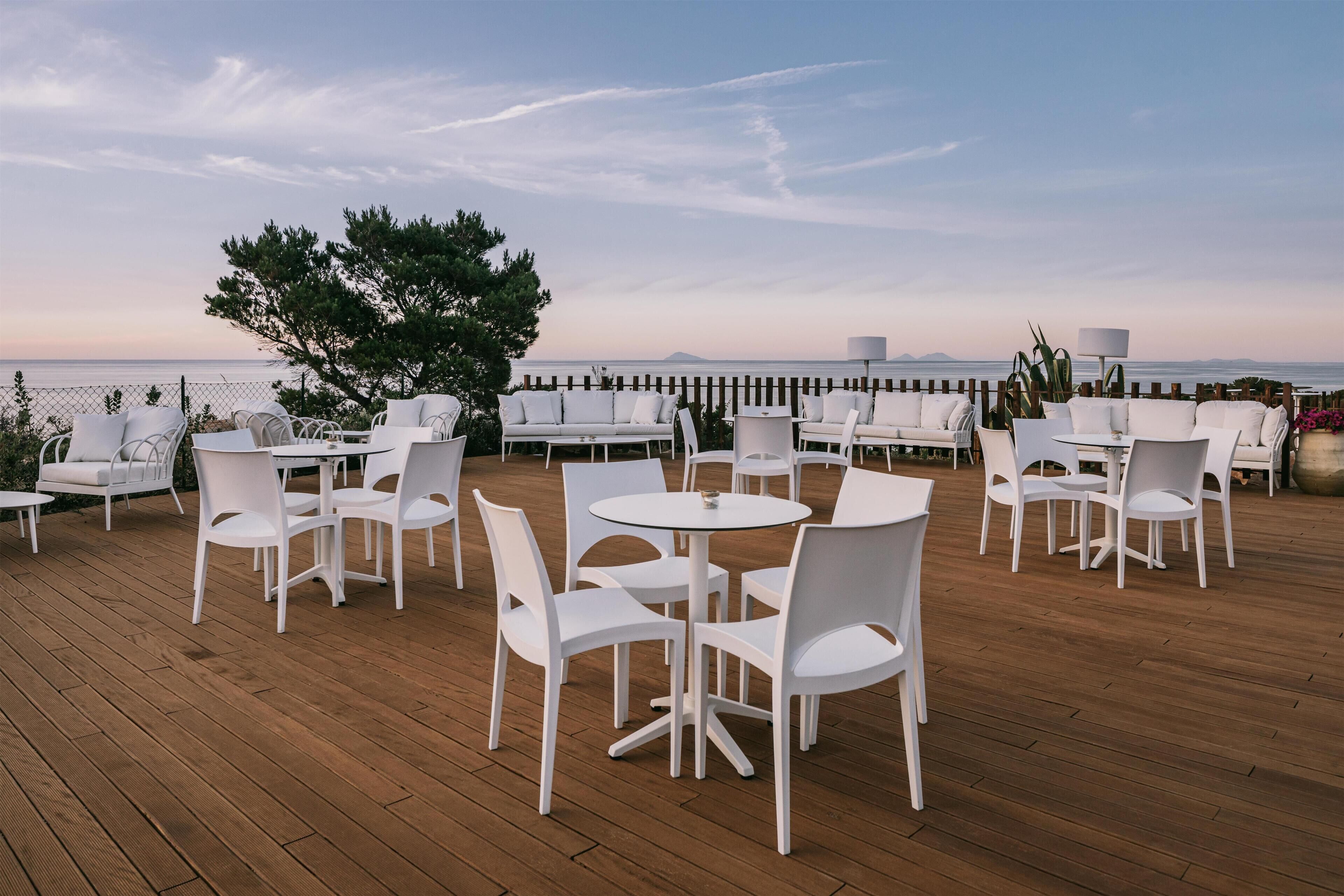 Una terraza con mobiliario exterior blanco, incluyendo mesas y sillas, con vista a un cuerpo de agua.