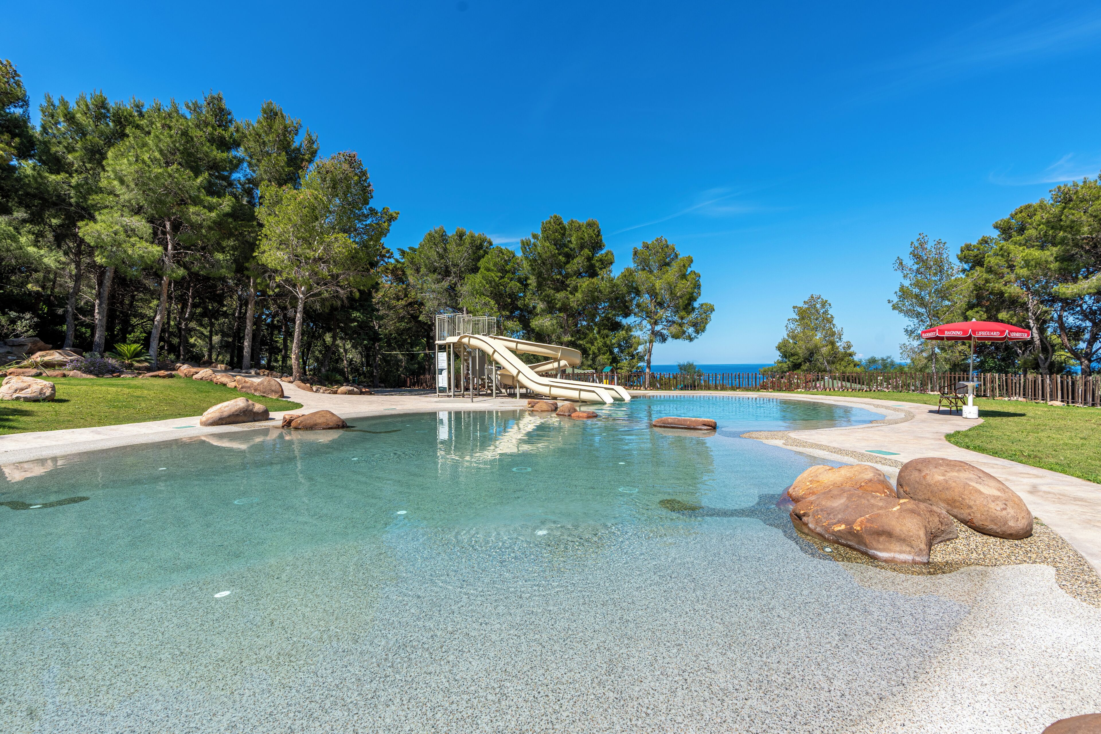 Una piscina cristalina con tobogán, rodeada de rocas y árboles, y una sombrilla roja al fondo.