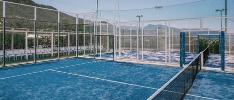 An indoor tennis court with a blue surface, surrounded by glass walls and metal fencing.