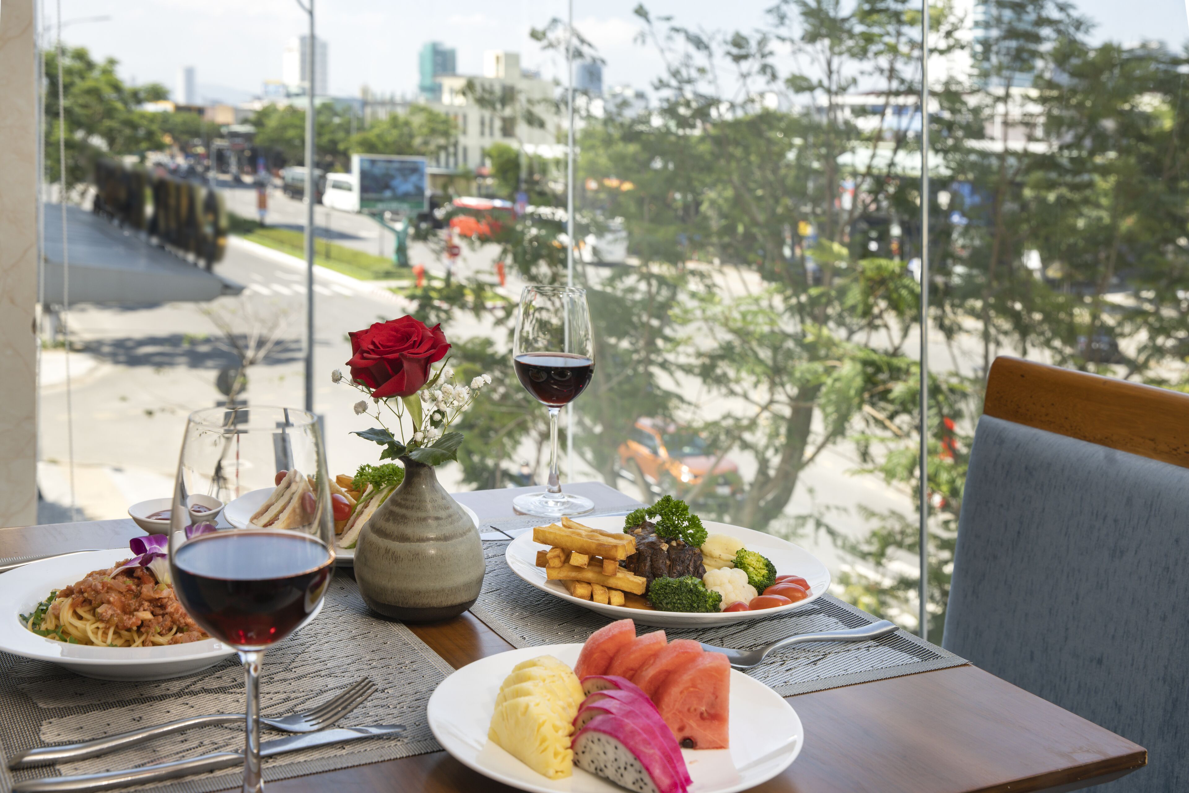A dining table with plates of food, a glass of red wine, and a vase with a red rose.