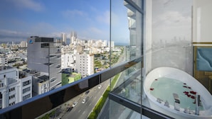 Une chambre d’hôtel située dans un immeuble de grande hauteur, offrant une vue sur la ville, dotée d’un balcon en verre et d’une baignoire moderne.
