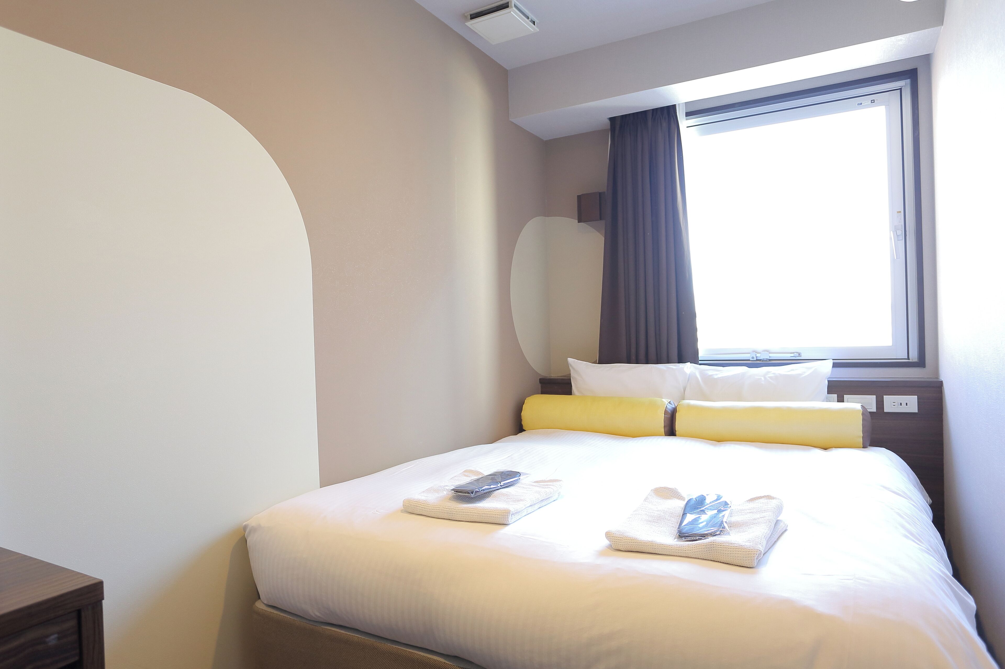 A hotel room with a neatly made bed, a bedside table, and a window with curtains.