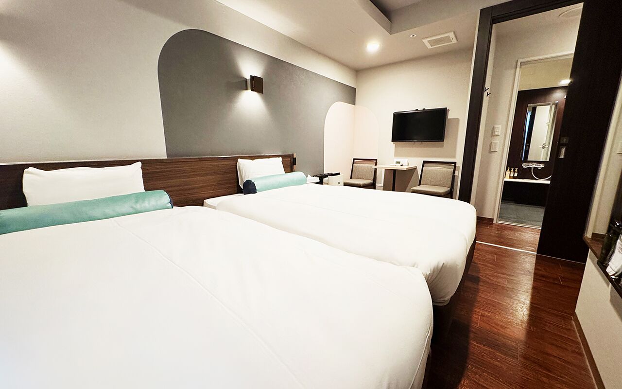 Ein Hotelzimmer mit zwei Betten, einem Schreibtisch, einem Stuhl, einem Fernseher und einem Spiegel.