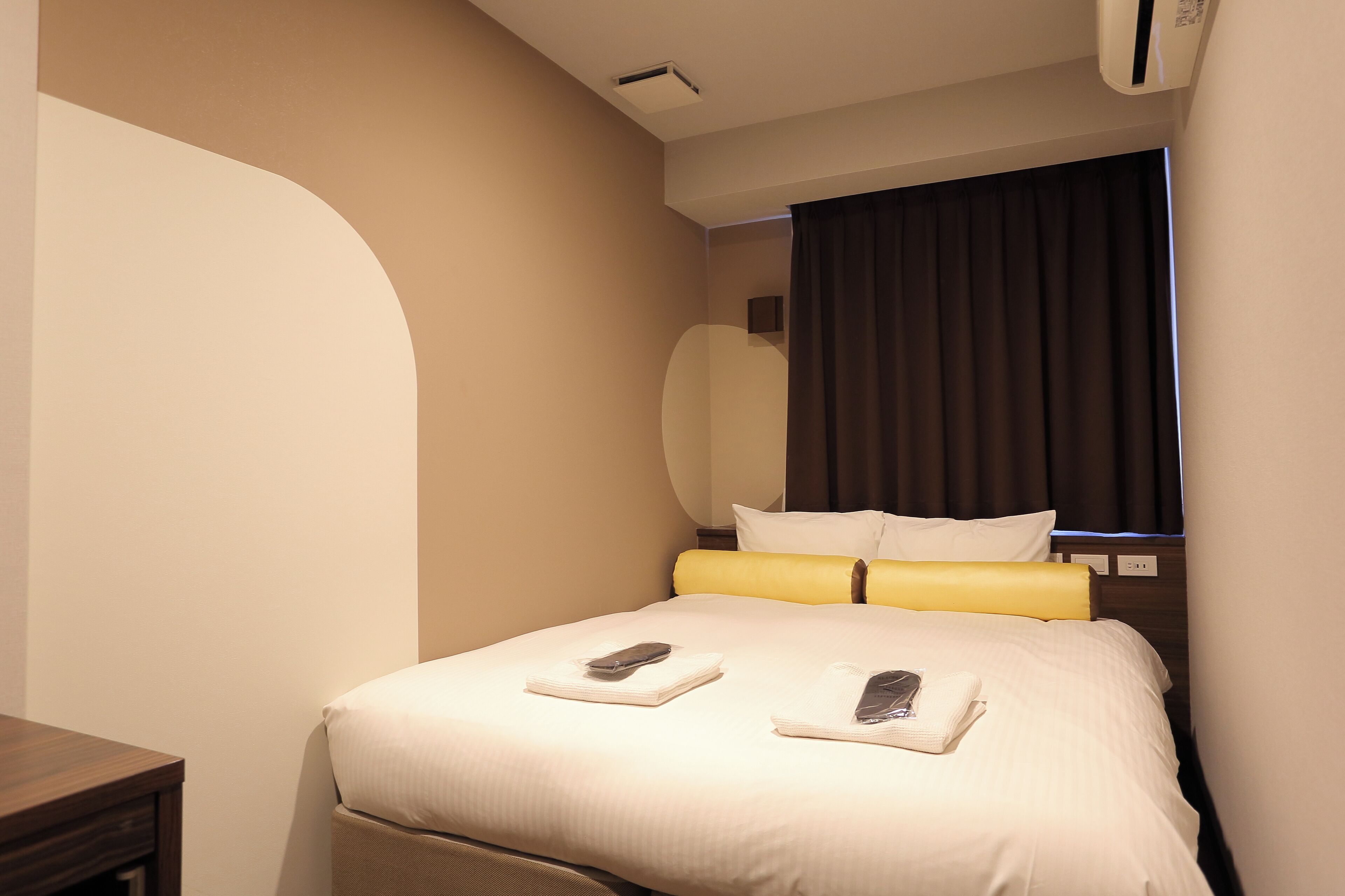 A hotel room with a bed, a bedside table, a window with curtains, and an air conditioning unit.