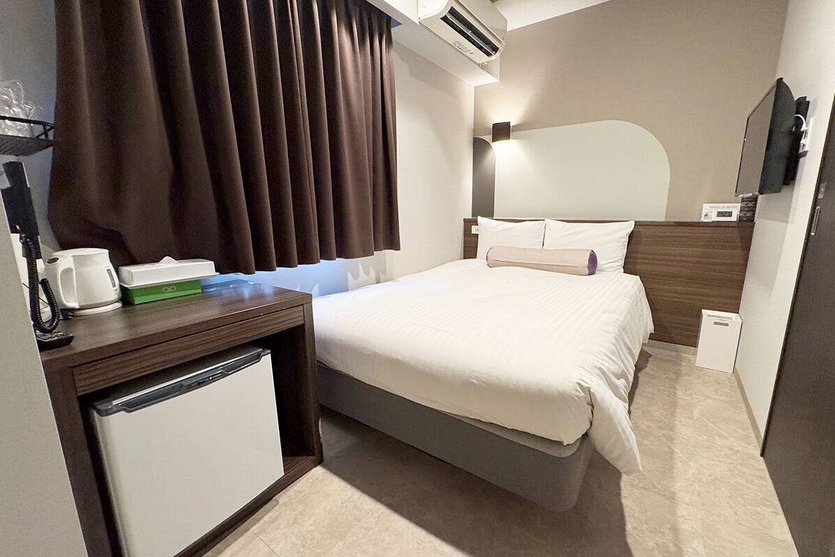 A hotel room with a bed, a desk with a coffee maker, a small refrigerator, a mirror, and a window with curtains.