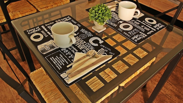 A glass-top table with a placemat featuring coffee-related graphics and text, a white mug, a small potted plant, and a set of cutlery. - Il Civico 2 (Bari)