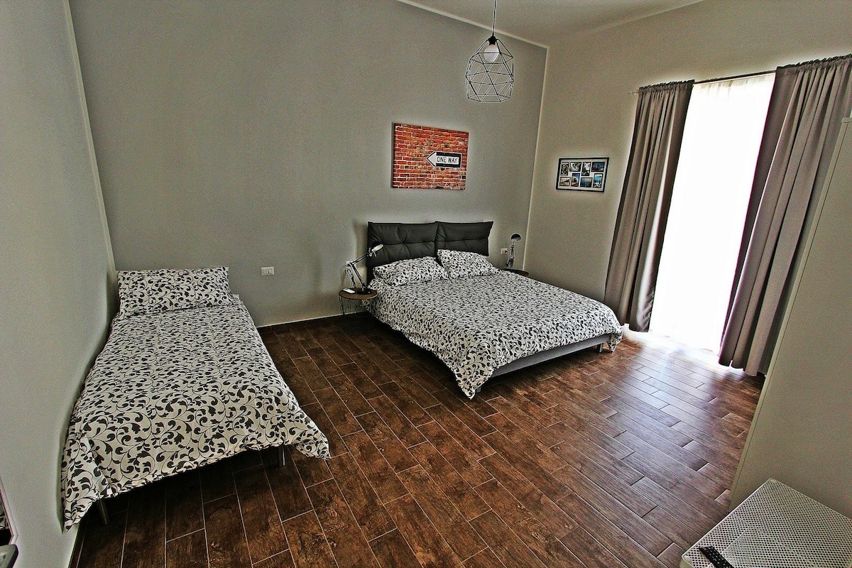 A room with two beds, a brick-patterned wall, a hanging light fixture, and a window with curtains.