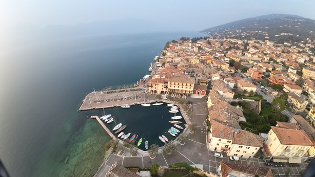 An aerial view of a coastal town with a marina, buildings with red-tiled roofs, and a clear view of the sea. - Hotel Garden (Torri del Benaco)