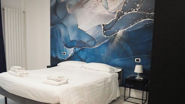 A neatly made bed with white linens and towels, a nightstand with a lamp, and a large abstract mural on the wall. -  La Boheme SMART Hotel (Turin)