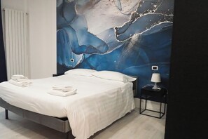A neatly made bed with white linens and towels, a nightstand with a lamp, and a large abstract mural on the wall. -  La Boheme SMART Hotel (Turin)