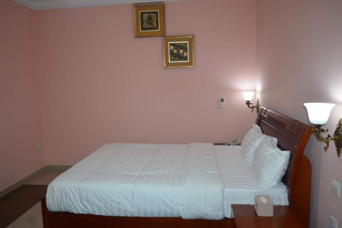 A neatly made bed in a room with pink walls, two framed pictures on the wall, and a bedside lamp.
