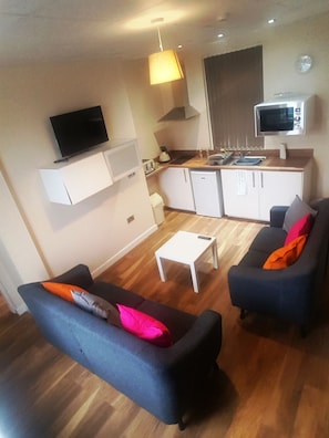A compact living space with a sofa, a coffee table, a TV mounted on the wall, a kitchenette with a microwave, and a dining area. - Premier Lodge (Wolverhampton)