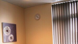 En stue med en mørkeblå sofa, røde og grå puder, et ur på væggen og et vindue med lodrette skodder.