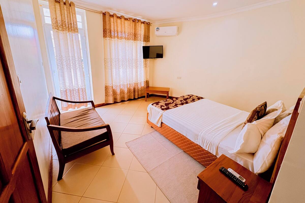 Habitación de hotel con cama, una silla, una mesita, televisión y ventana con cortinas.