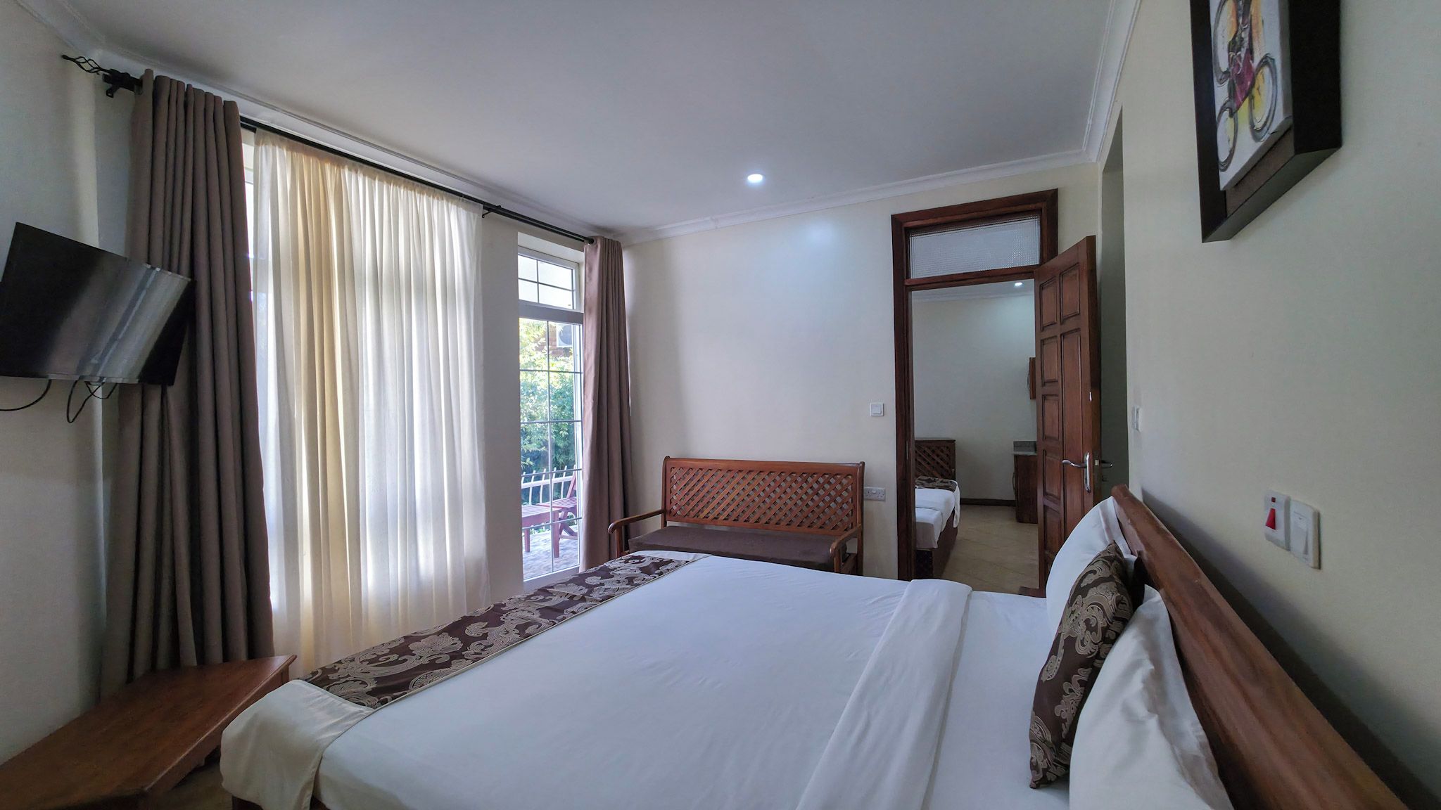 Habitación de hotel con cama, televisor montado en la pared, banca de madera, puerta y ventana con cortinas.