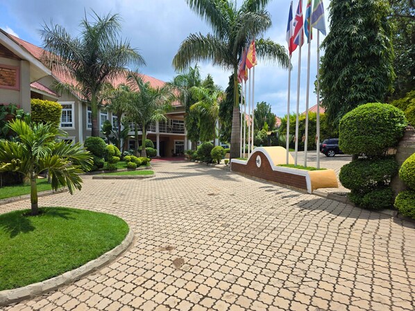 A paved entrance with a curved wall, palm trees, and flags. - Masailand Safari & Lodge (Arusha)