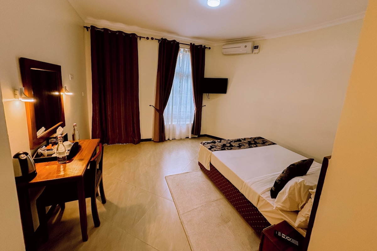 A hotel room with a bed, a desk, a chair, a mirror, a window with curtains, an air conditioning unit, and a television mounted on the wall.